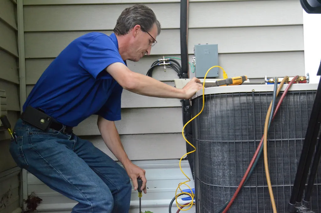 An HVAC technician in a polo shirt servicing a residential outdoor condenser unit beside a house, with gauges and tools. An HVAC technician in a polo shirt servicing a residential outdoor condenser unit beside a house, with gauges and tools.